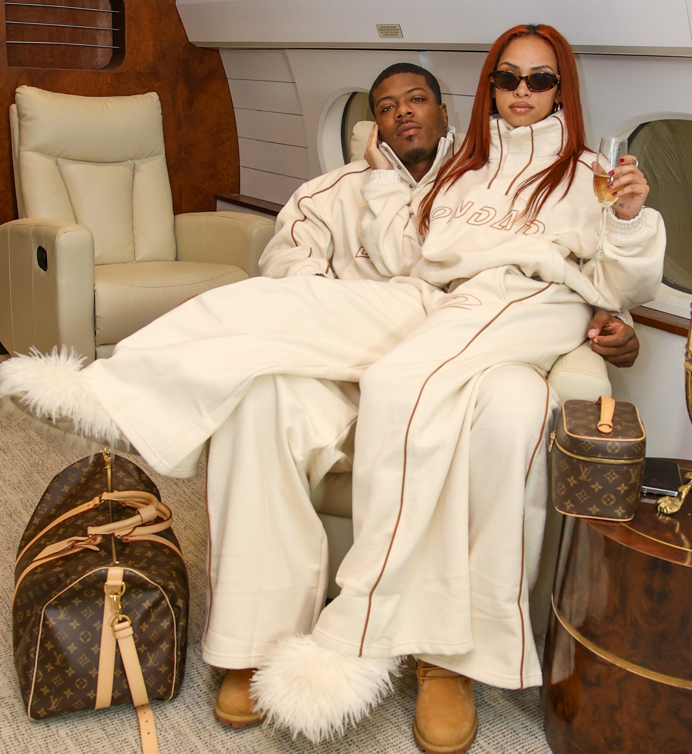 Two people in white outfits sitting on a private jet with Louis Vuitton bags.
