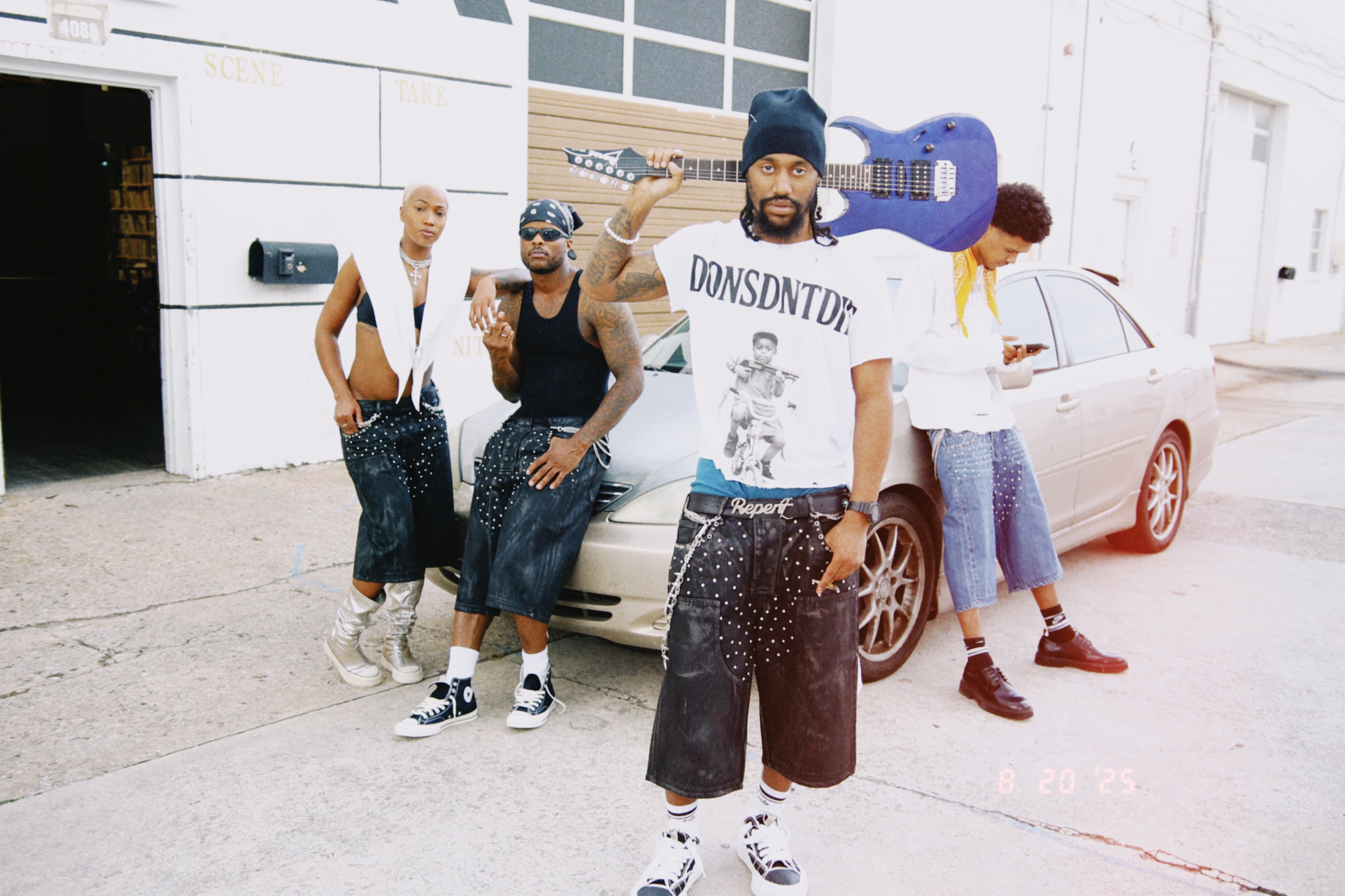 Group of models wearing DonsDntDie jorts and tees posing in front of a white building and vintage car, showcasing streetwear fashion from Don Dada Official.